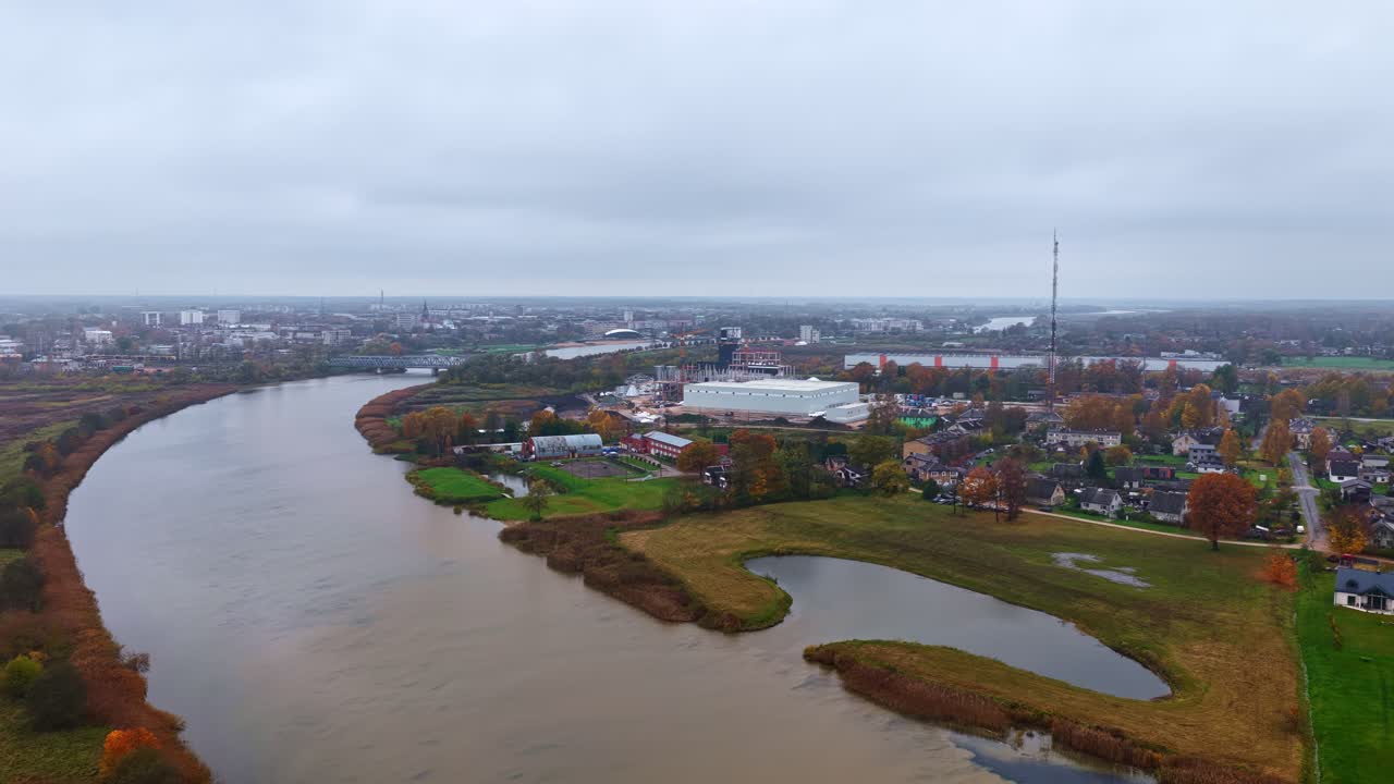 A Calm River Winds Through a Peaceful Town Surrounded by Autumn Trees Under a Cloudy Sky - Aerial Drone Shot