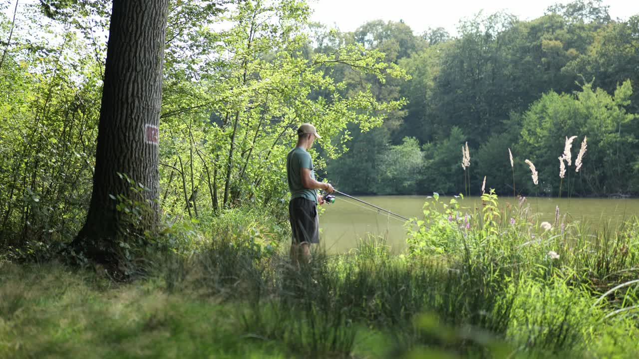 Giovane pescatore al bordo di un lago torbido circondato da alberi gira il mulinello e recupera la lenza, ripresa in avvicinamento
