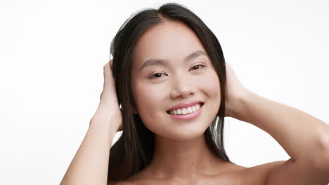 a beautiful young woman with long dark hair smiling and touching her hair, looking at the camera on a white background