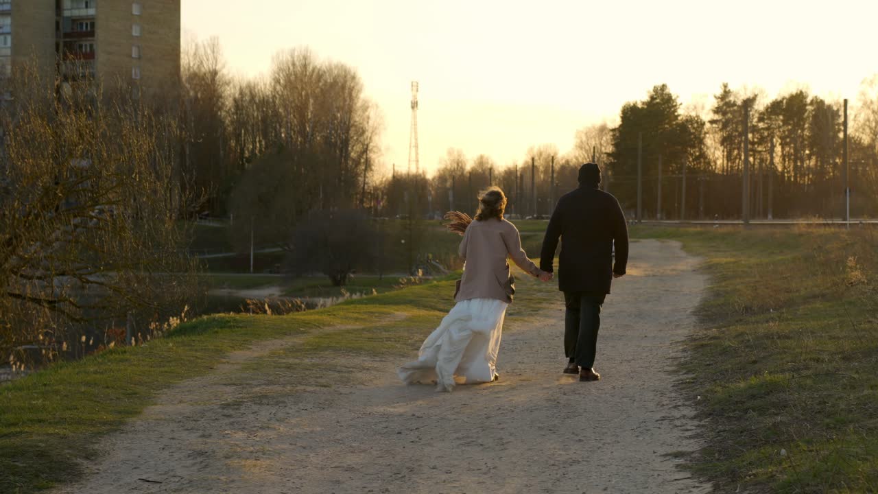 Newlyweds holding hands, strolling along winding path during golden sunset, bride's gown flowing softly, capturing intimate moment of love and tender connection
