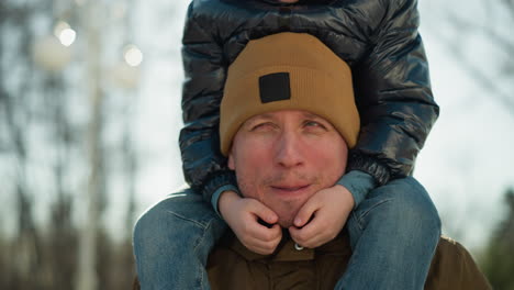 close-up of a father carrying a child on his shoulders, smiling and talking, with the child's hands resting on the father's face, light rays are reflecting from behind