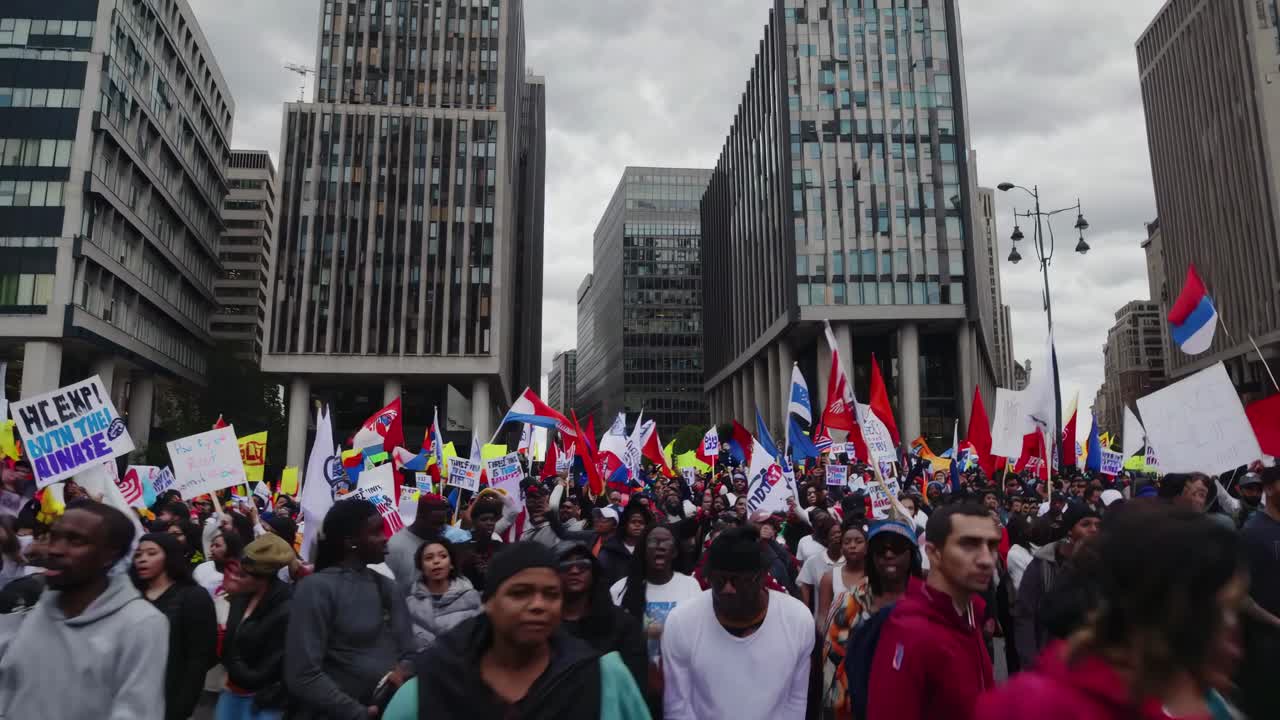 Wide-angle video shot of a diverse crowd at a protest, with skyscrapers in the background