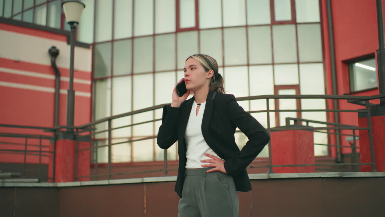 Young woman in formal attire talking on phone near fence in city with concerned expression, appearing worried and tense, standing against backdrop of red and glass building structure in urban setting