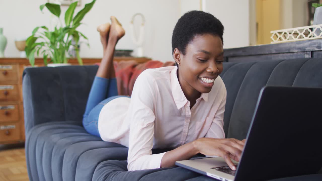 Happy african american woman lying on sofa in living room, using laptop