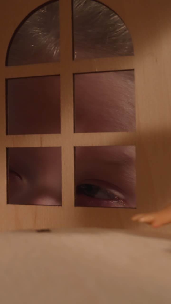 Blonde toddler girl with fever peeks into window of handmade wooden dollhouse with pretty dolly at small round table close view zoom in