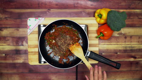 una vista superior de la adición de chile triturado a los ingredientes en la sartén, colocados en una estufa, dos grandes pimientos amarillos y rojos y un brócoli verde sobre la mesa