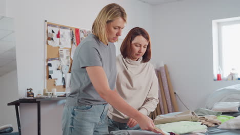 two dress makers attentively pick up textile fabric while conversing at busy worktable covered with fabric pieces, patterns, and color swatches, inside bright fashion workshop with large window