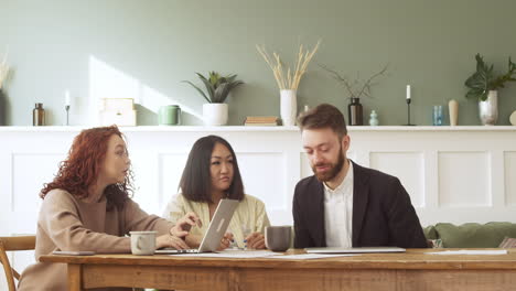 group of three multiethnic colleagues sitting at table, talking together and working on laptop computer