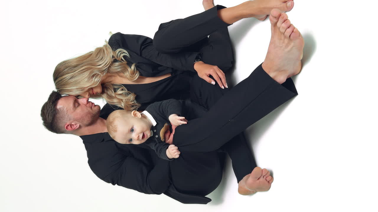 Couple in love wearing black suits sit close to each other on the floor. Happy parents of an infant child in studio. Vertical screen.
