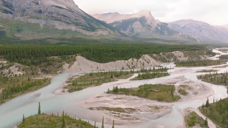 órbita-Aérea-De-Los-Arroyos-Fluviales-Que-Fluyen-Entre-Bosques-De-Pinos,-Montañas-Rocosas-Canadienses-En-El-Fondo,-Parque-Nacional-De-Banff,-Alberta,-Canadá