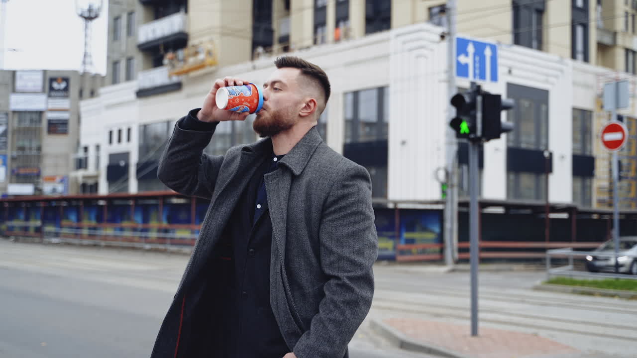 Charming young businessman dressed in suit and coat drinking coffee. Man outside a modern building.
