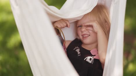 a little smiling girl sits in the hammock holding a gift bag with a present in her hands