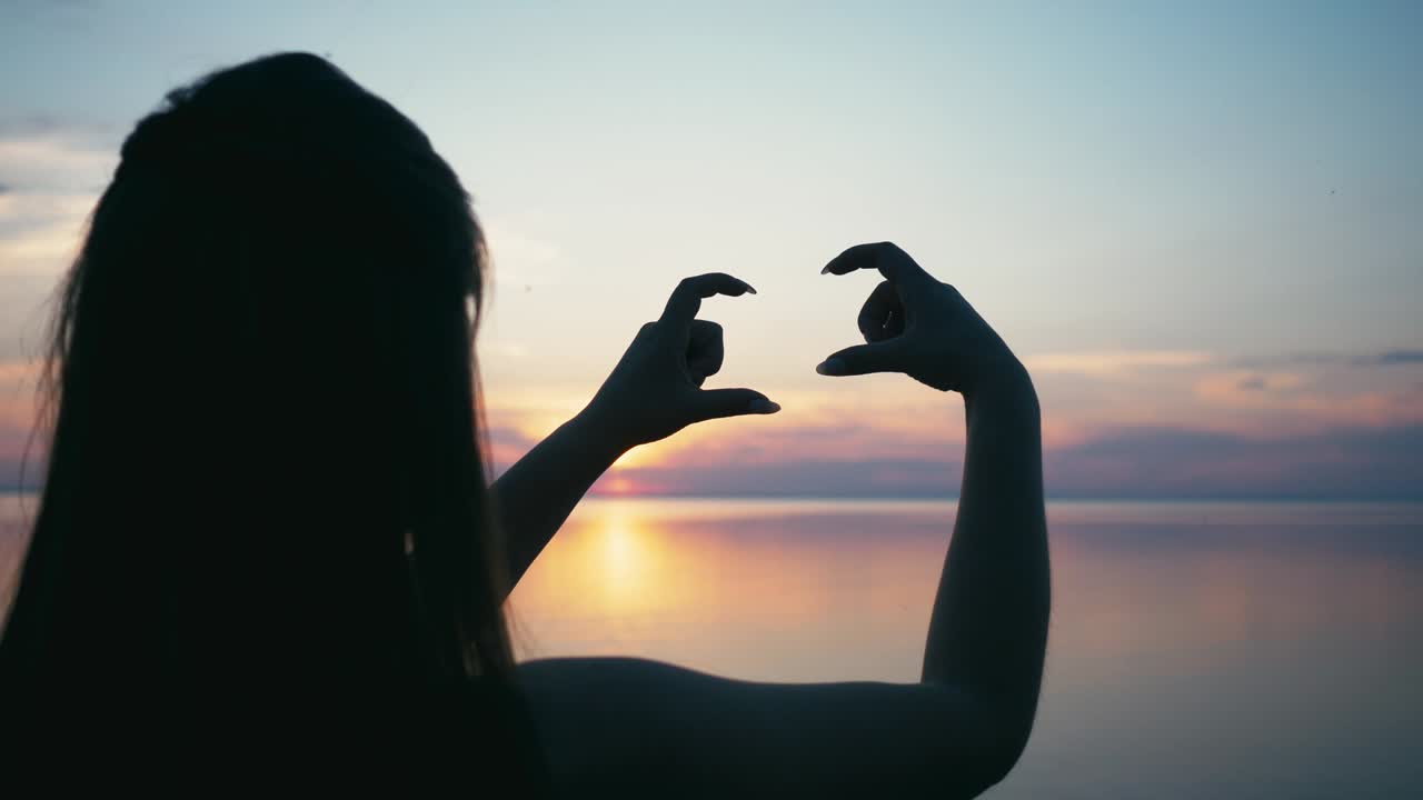Beautiful Woman making heart shape with hands at sunset Girl holding up love symbol gesture with orange sun flare