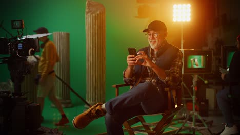 prominent successful director sitting in a chair on a break using smartphone. on the studio film set with high-end equipment professional crew shooting high budget movie. zoom in looking at camera