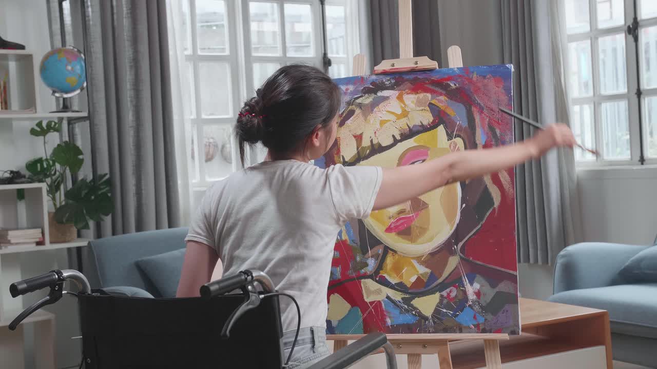 Back View Of Talented Female Artist Sitting On Wheelchair, Enthusiastically Using Paint Brush She Creates With Oil Painting. Creative Studio With Canvas
