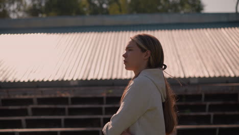 girl in hoodie with long hair tied back walking outdoors, looking pensive touched her cheek as her hair gently flutters in breeze, with roof and trees in background