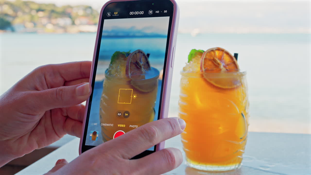 Close up of a woman's hand filming an orange cocktail on her phone with a blurred view of the sea on the background