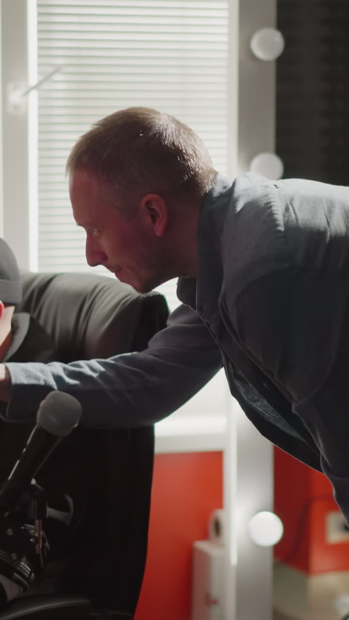 dos hombres están en una sala de estudio, uno sentado frente a una computadora, probablemente un productor, con ropa negra, mientras que el otro, vestido con una camisa de manga larga, se inclina, explicando algo atentamente