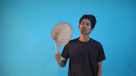 smiling young indian boy relaxing in home with hand made jute fan in blue color background studio