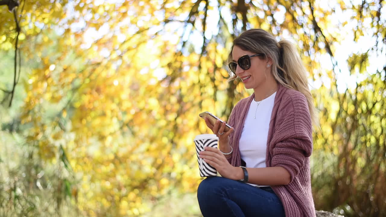 Blond kvinde, der skriver på sin telefon i parken, mens hun drikker kaffe