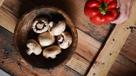 hand putting a red pepper next to a wooden bowl with white mushrooms inside on rustic wooden texture as background-flat lay,steady cam