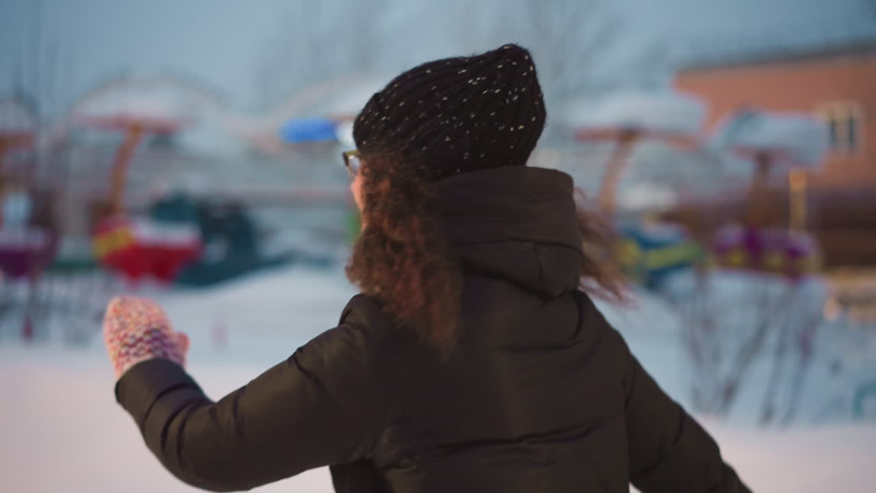 Female skater in black winter jacket and knit hat with snowflakes skating outdoors during cold evening near snowy amusement park rides and glowing streetlights, enjoying peaceful winter moment