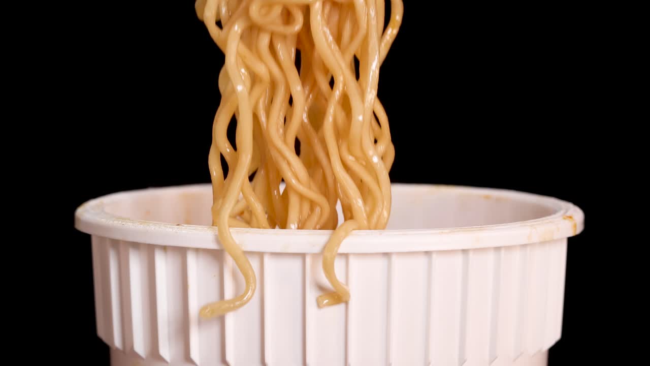 A hand skillfully lifts cooked instant noodles from a white cup using chopsticks against a black background, with even studio lighting and close-up framing