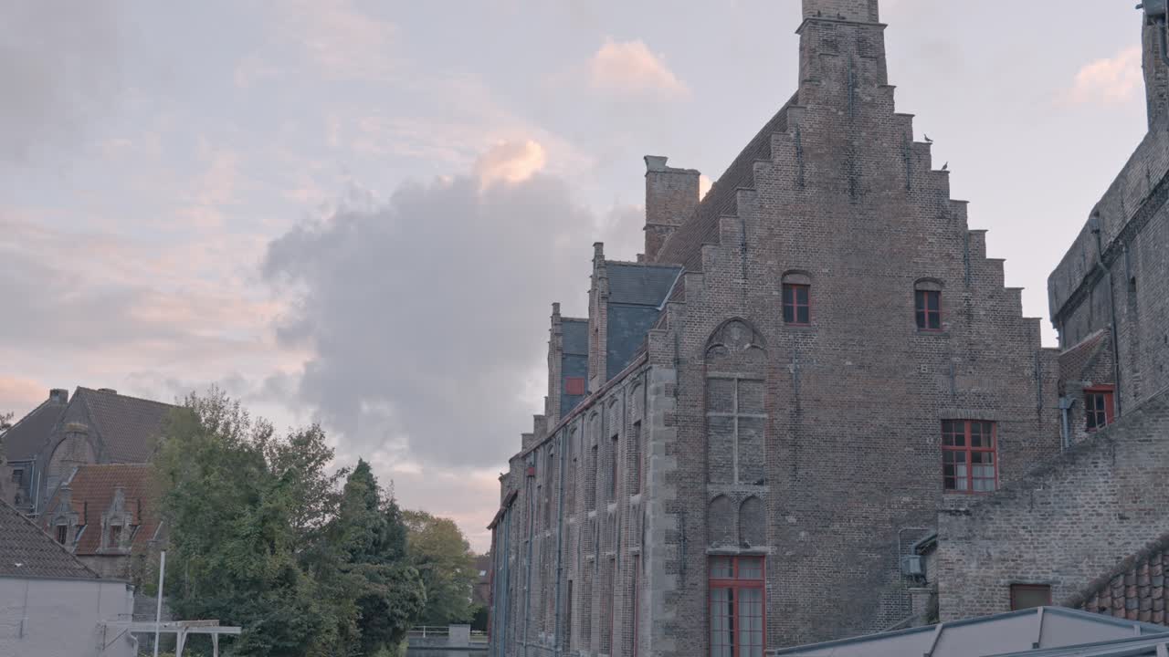 An atmospheric view at dusk in Bruges, Belgium, capturing a tranquil canal flanked by the imposing Gothic brick architecture of the Sint-Janshospitaal (Old St. John's Hospital)