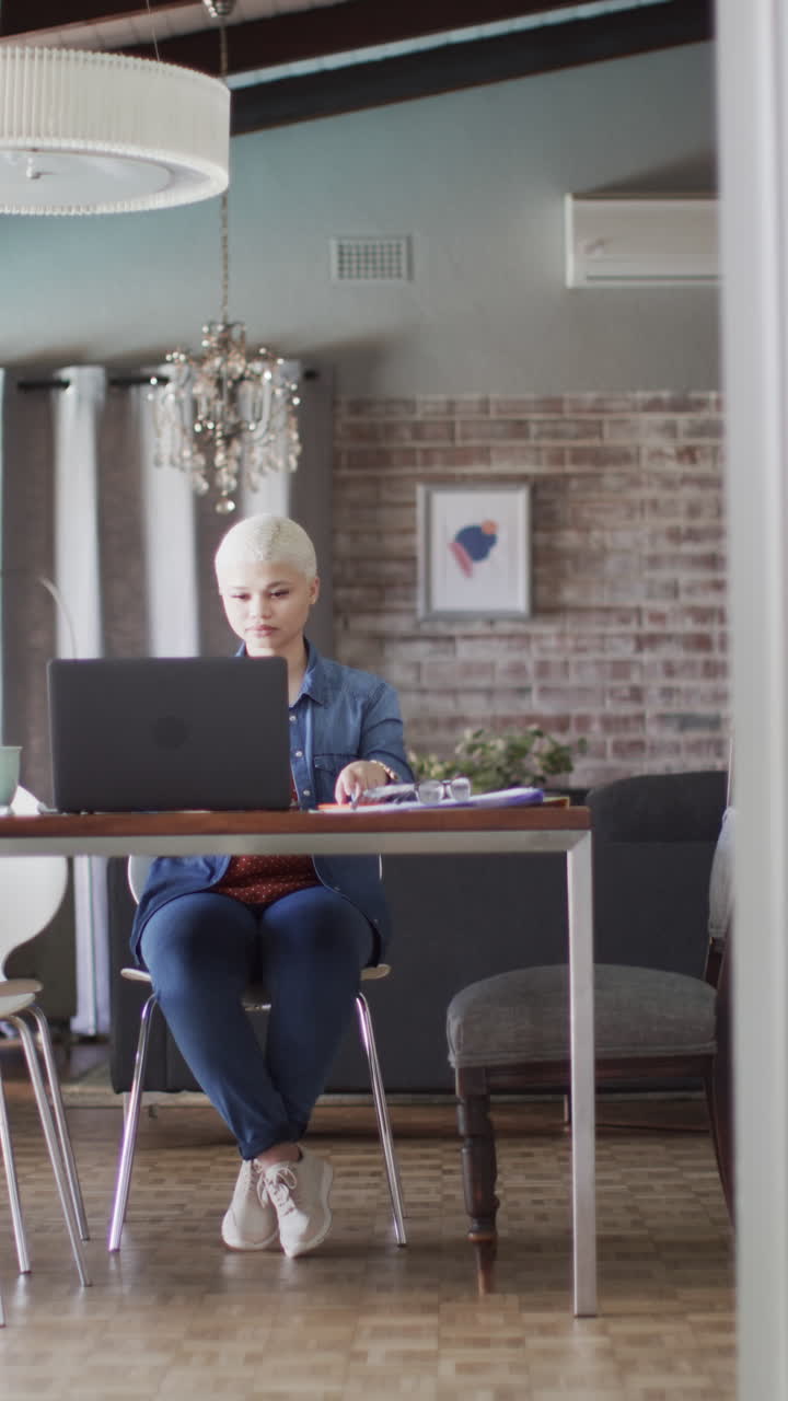 Vertical video of busy biracial woman working with laptop at home in slow motion