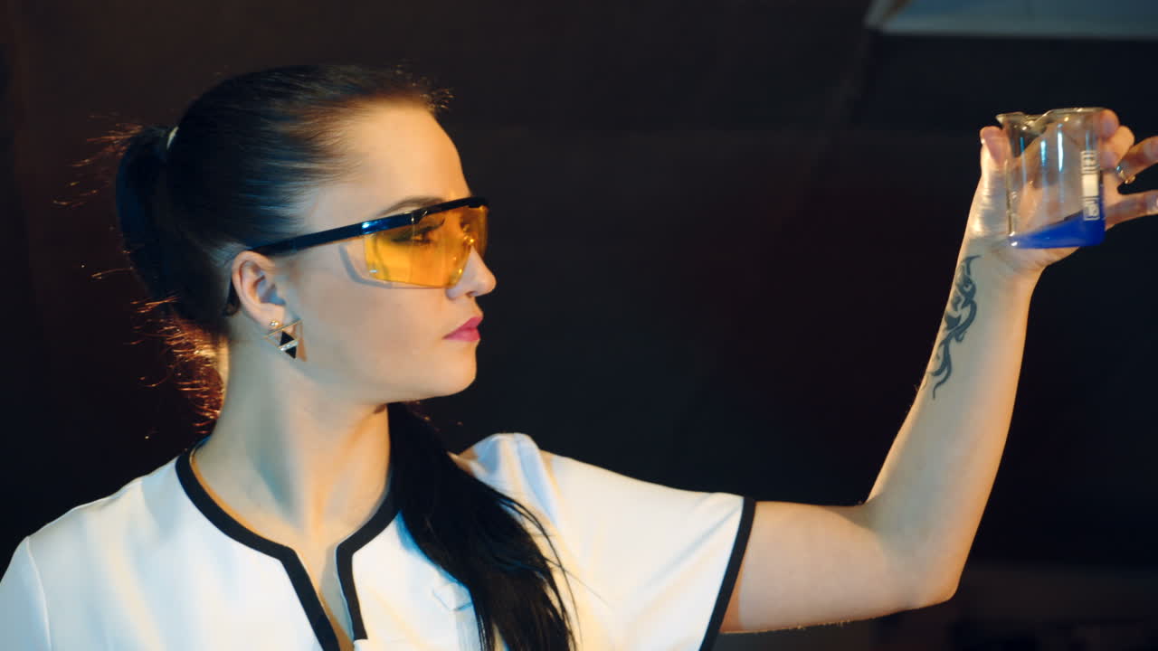 A woman in protective glasses is conducting experiments in the laboratory. Chemist is holding a jar of blue liquid and looking the reaction on the black background of the room