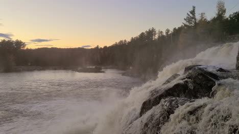 sliding drone as mist sprays from ledge waterfall that plunges into a lake at dusk