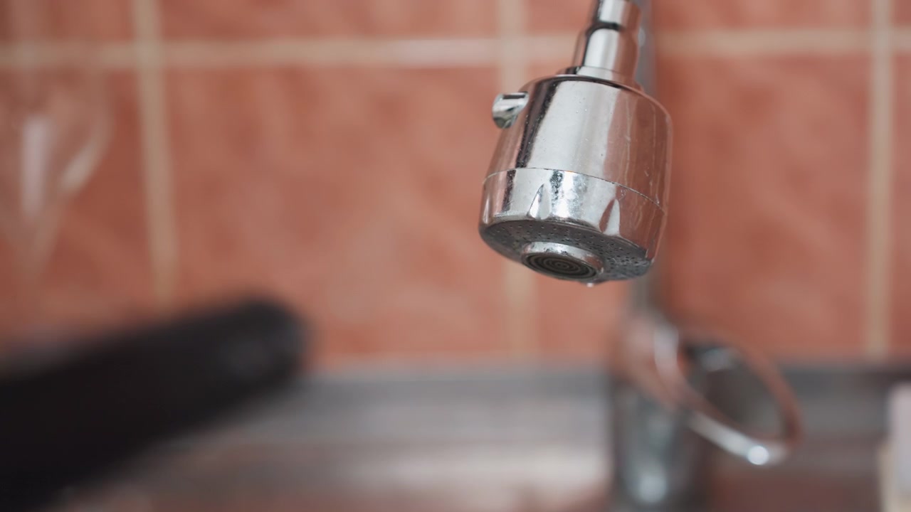 Primo piano di un rubinetto da cucina in una scena di pulizia domestica, soffione cromato con goccioline d'acqua, paraschizzi piastrellato, bordo del lavello, spugna e attrezzo sfocati sul bancone, attenzione sui depositi minerali e sulla finitura usurata, atmosfera tranquilla