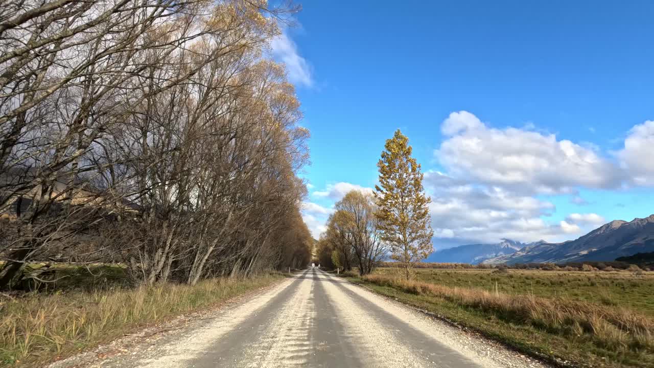 Forward-moving car view along rural gravel road, autumn trees, dramatic clouds, and distant mountains