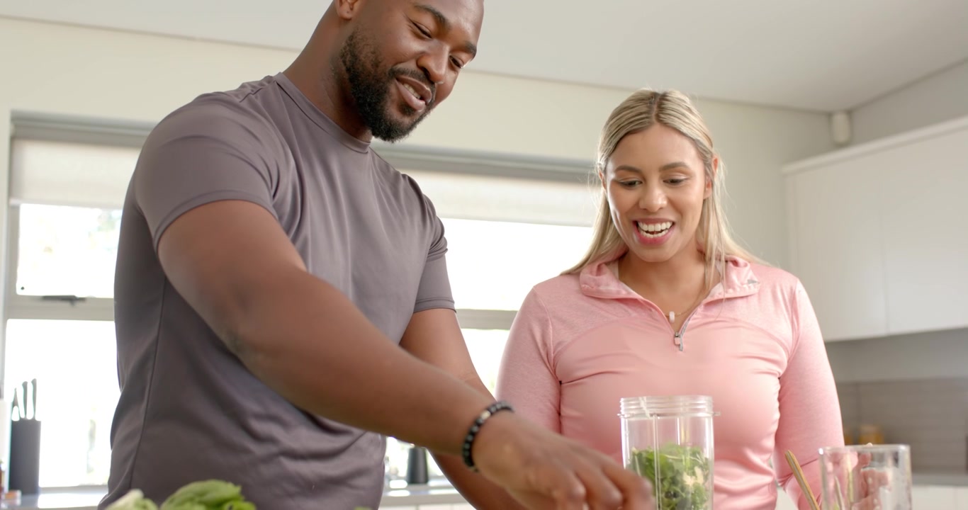 Casal preparando bebida em ilha de cozinha, homem pegando vegetais, adicionando limão no liquidificador, mulher sorrindo.
