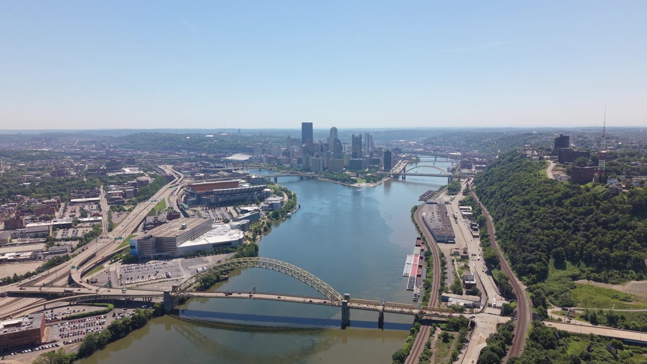 Prospettiva aerea da drone che segue un ampio fiume verso un grande ponte autostradale e lo skyline urbano del centro città in una giornata limpida e soleggiata
