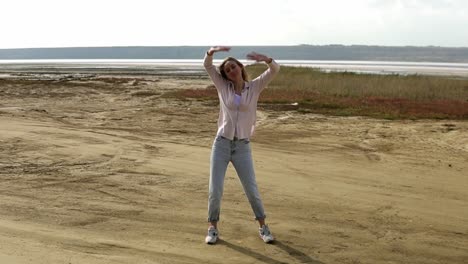 carefree young woman is dancing alone standing on sand beach with calm lake on background