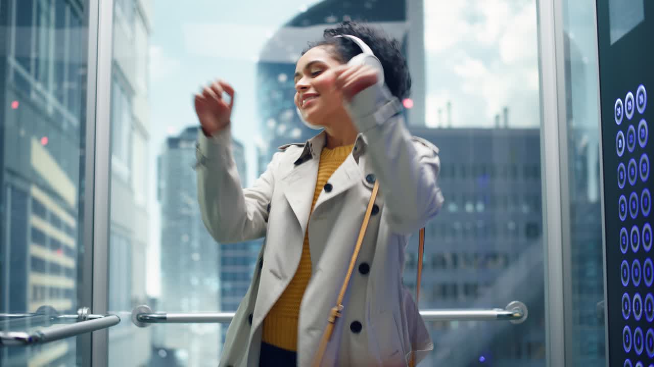 현대 비즈니스 센터의 사무실에 유리 엘리베이터를 타고있는 무관심한 흑인 여성 아름다운 라틴 여성은 행복하고 미소 짓고 헤드폰으로 음악을 듣고 엘리베터에서 춤을 추고 있습니다.