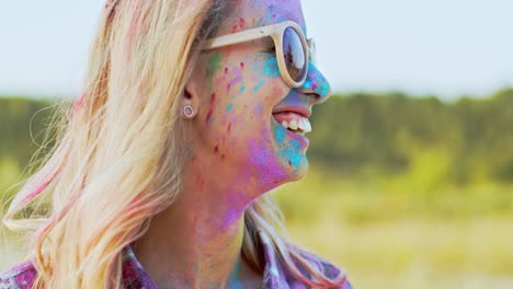 gros plan de la belle jeune femme blonde à lunettes de soleil souriant et regardant sur le côté tandis que son visage est dans des peintures colorées alors qu'elle célèbre holi, vacances