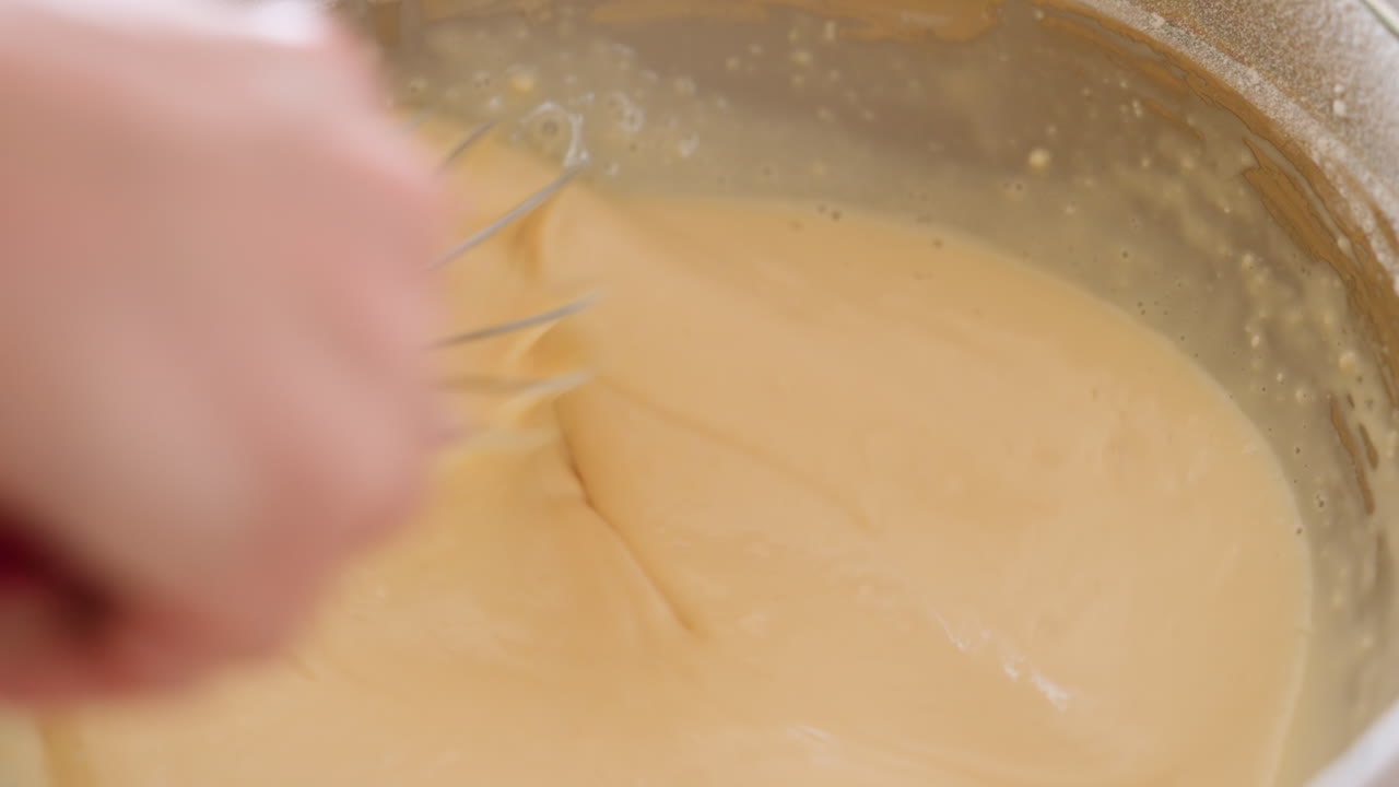 Close up of baking process as chef stirs creamy batter in stainless bowl using whisk, showing texture and movement during homemade cooking preparation in kitchen environment