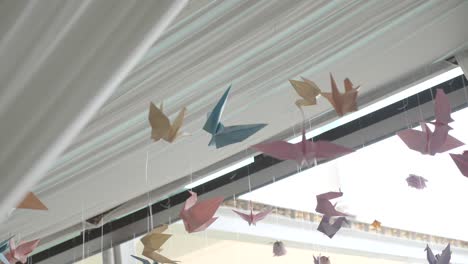 wider shot of lots colourful pastel origami paper crane birds floating in breeze, hung from roof, blue, yellow, pink and orange