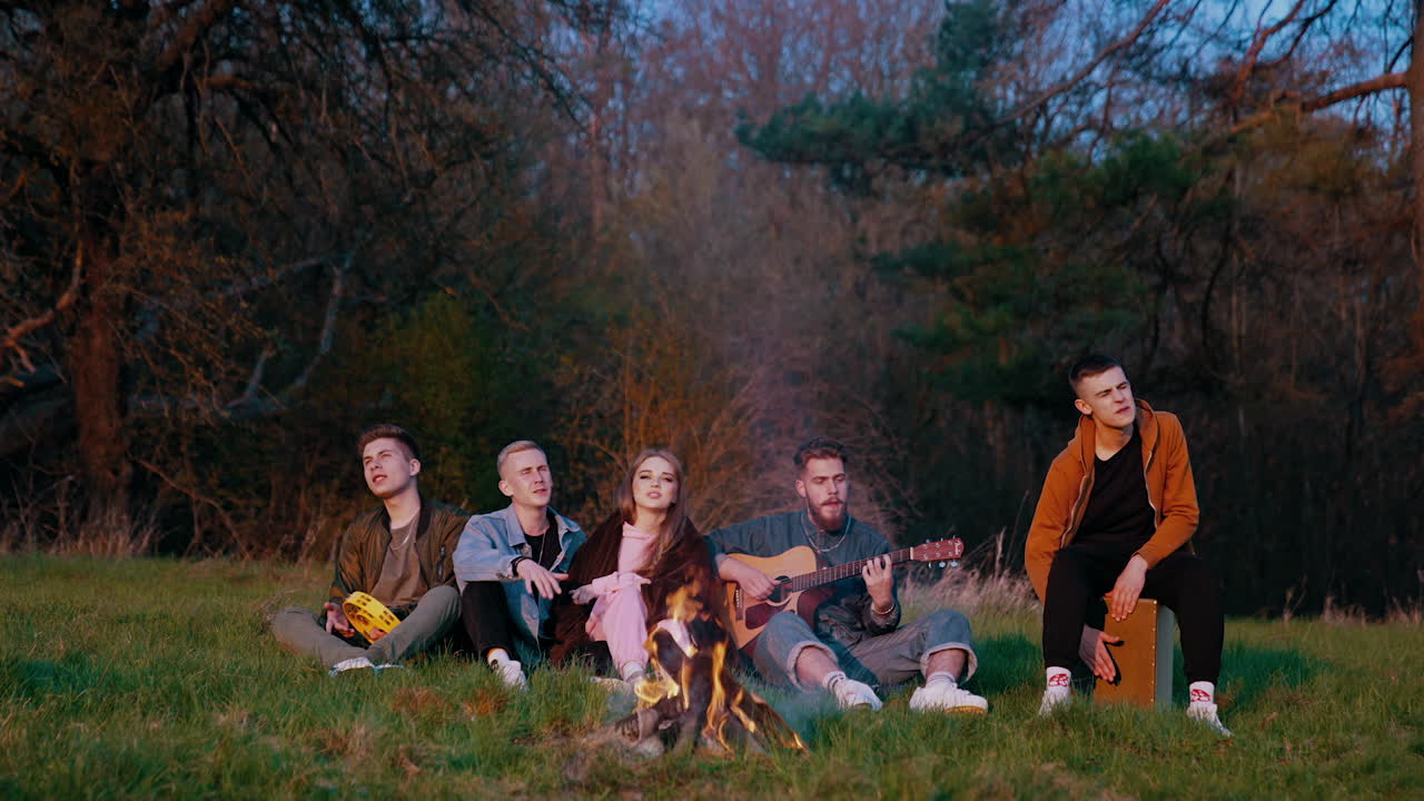Young people sitting by the fire. Friends singing together to the guitar while sitting near bonfire in summer. Young men and beautiful girl rest in nature in the evening.
