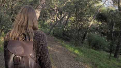 slow motion following the back of young blonde woman carrying a backpack while walking among pine trees on a path