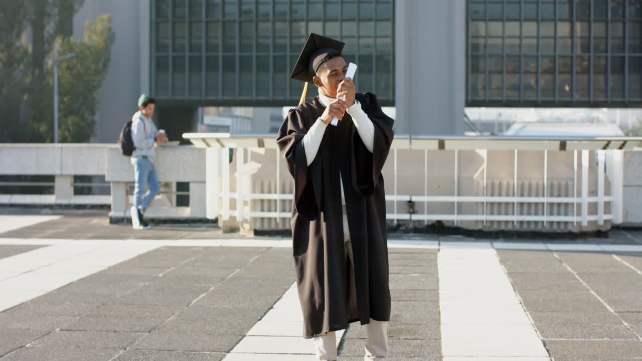Uomo afroamericano adulto che tiene un diploma arrotolato, lo bacia e lo solleva, festeggiando sulla terrazza.