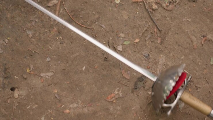 Close up of sword resting on dry soil scattered with leaves and twigs. metallic blade reflects light while hilt features red fabric detail emphasizing texture against earthy background