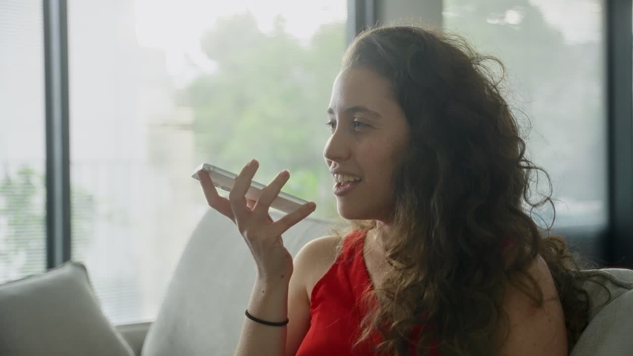 Woman recording voice message on mobile phone while sitting on sofa in her apartment. Cheerful and happy she is leaving an audio sms to friend. Business or gossip or telling interesting story