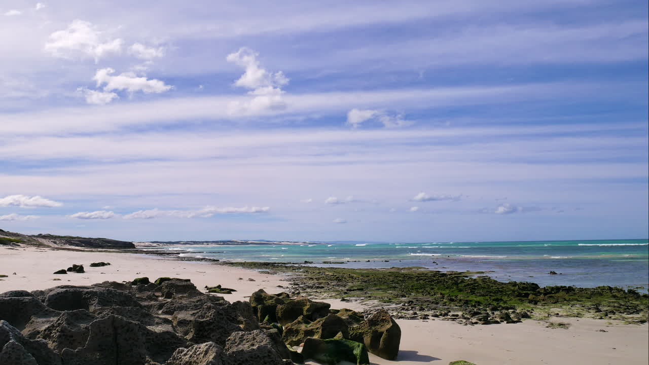 timelapse - nubes moviéndose sobre una playa aislada blanca con olas rodando, rocas negras escarpadas en primer plano, día soleado