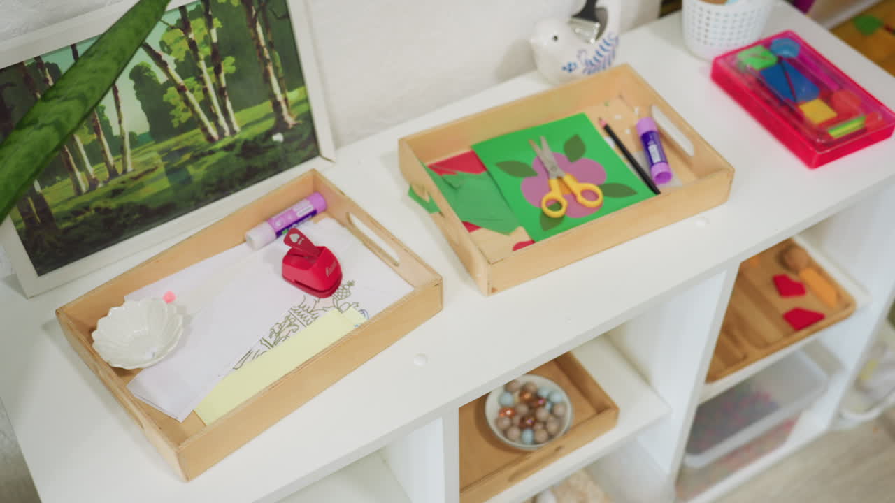 Organized kindergarten craft trays containing colored paper, child scissors, glue stick, crayon, punch, and printed drawing sheet placed neatly beside painting and snake plant in classroom