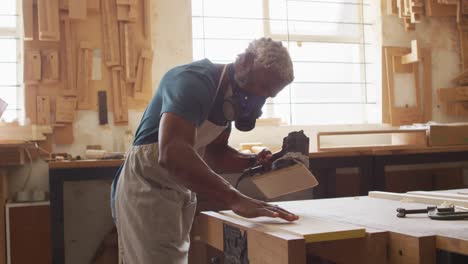 african american male carpenter wearing protective mask using electric grinder to grind wooden plank