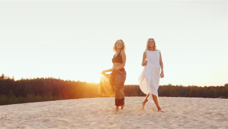 Dos-Mujeres-Jóvenes-En-Hermosos-Vestidos-Están-En-La-Playa-Al-Atardecer,-El-Viento-Juega-Un-Hermoso-Vestido-Estable