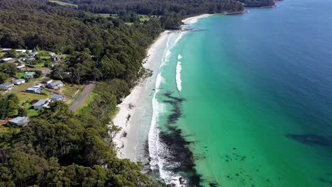 Aerial-drone-pan-over-white-sandy-beach-as-slow-moody-waves-roll-in-on-beautiful-summers-day,-with-small-town-and-forest-road-in-distance